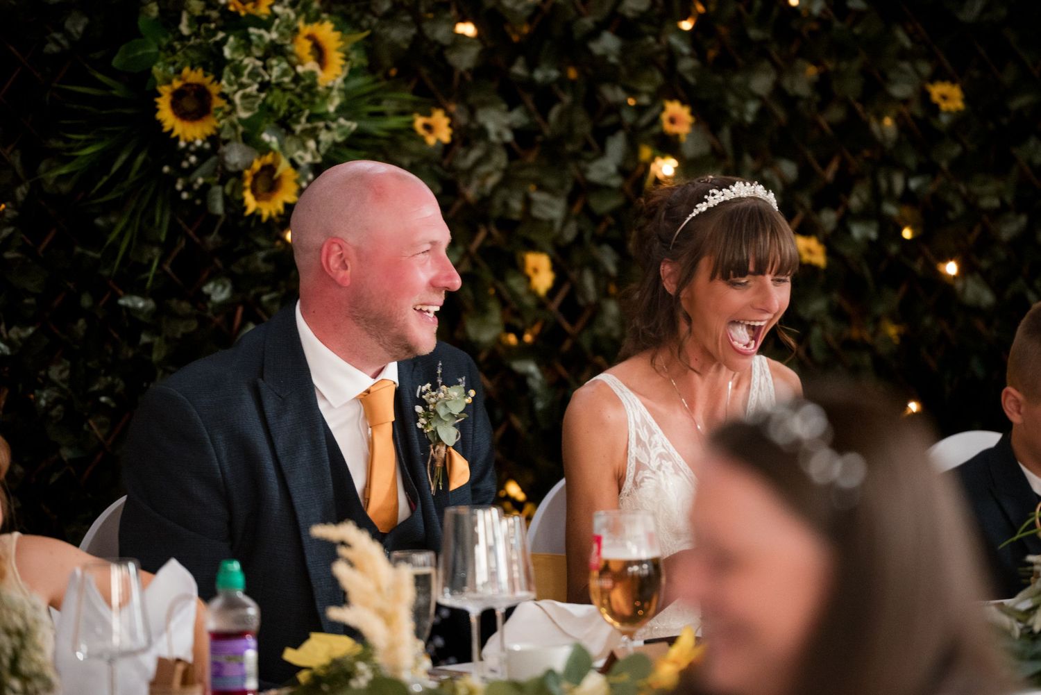Wedding reception guests share joyful laughter during dinner at an elegantly decorated table with floral arrangements.