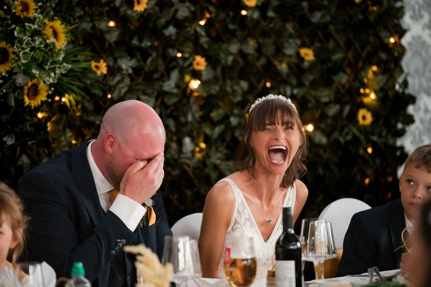 Wedding guests laughing and enjoying themselves at an evening reception with glowing lights in the background.
