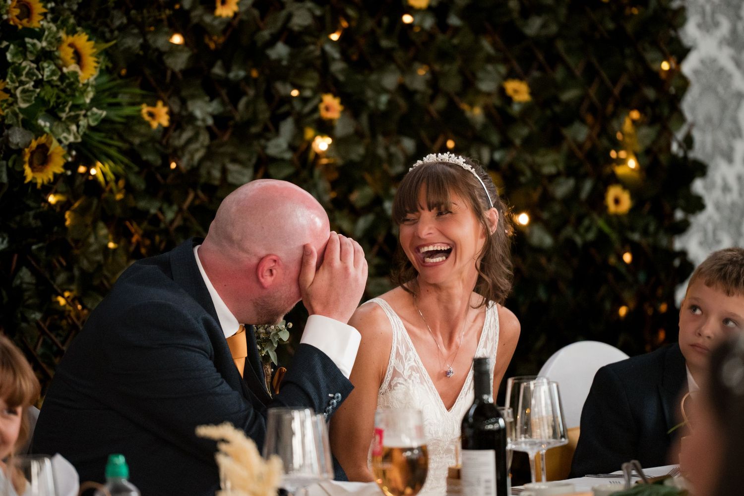 Candid moment of laughter during wedding reception dinner with wine bottles and glowing lights in background.