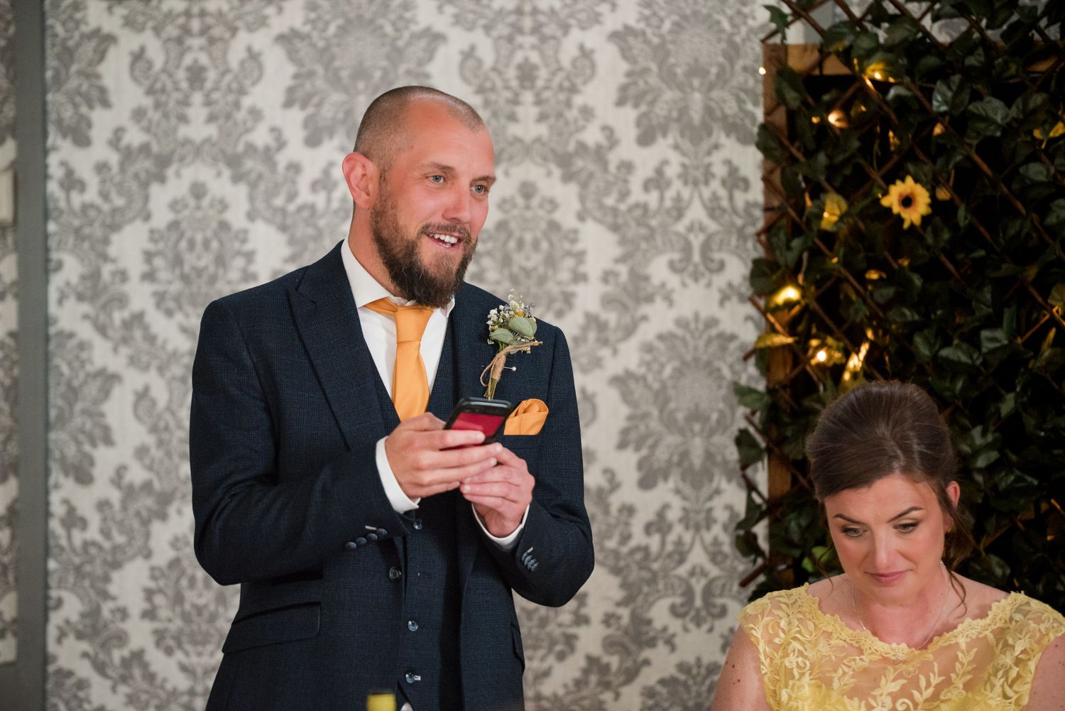 Man in dark suit delivers speech while holding paper at formal event with decorative wallpaper backdrop.