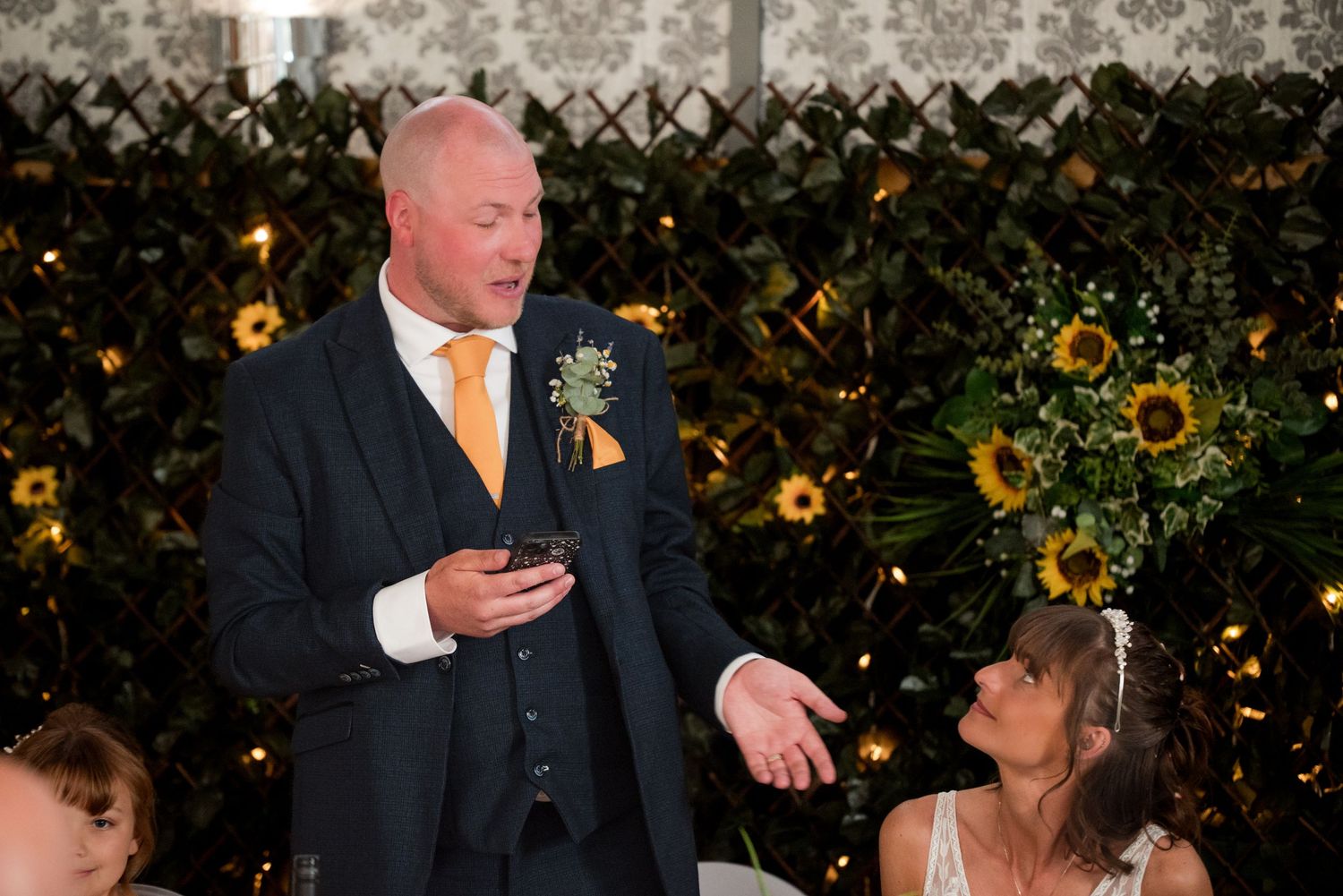 A couple shares a romantic dance surrounded by a wall of sunflowers and twinkling lights at an outdoor evening event.
