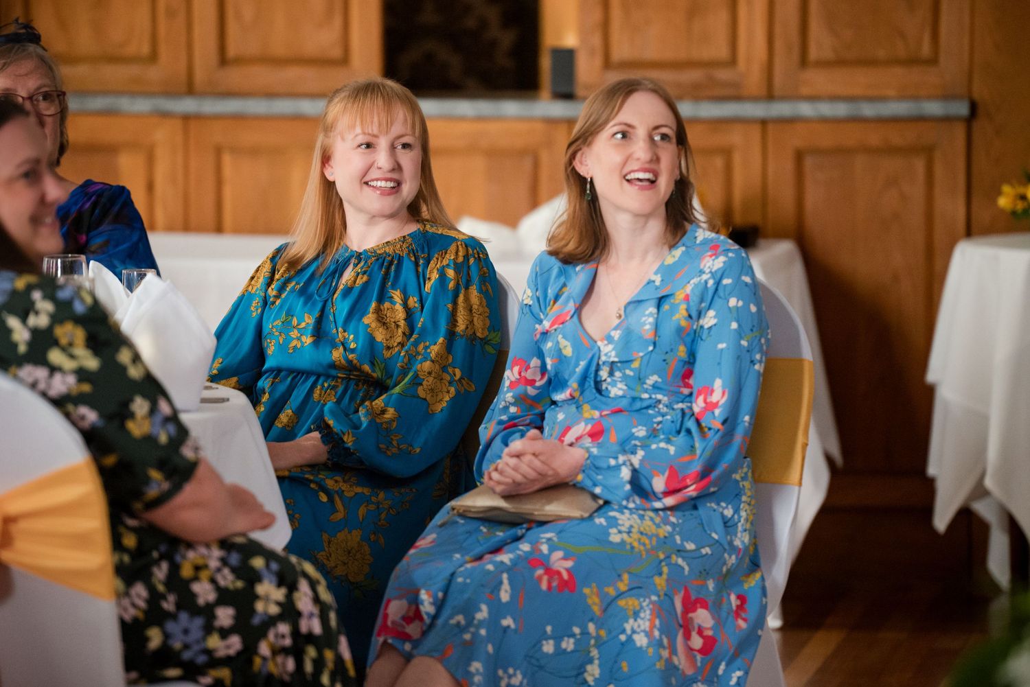 Wedding guests in colorful floral dresses share joyful laughter during a reception celebration.