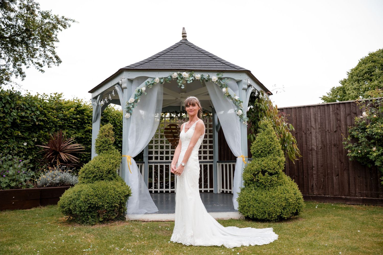 Elegant white gazebo decorated with sheer curtains and surrounded by manicured shrubs in a garden setting.