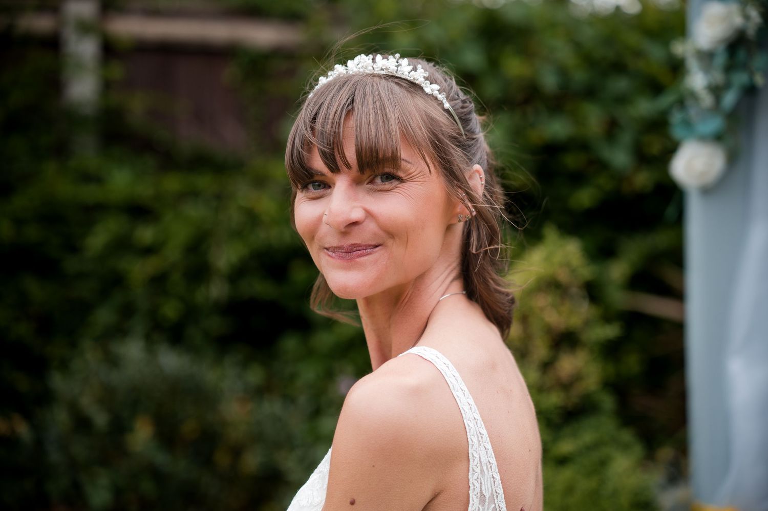 A person in a white dress smiles while posing in front of lush green foliage outdoors.
