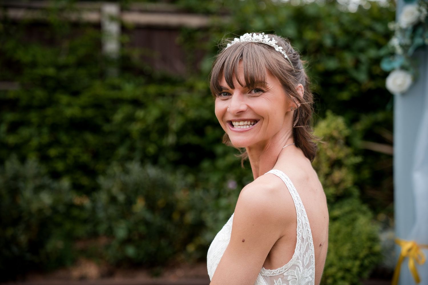 A bride in a white lace wedding dress smiles joyfully while standing in a garden setting.