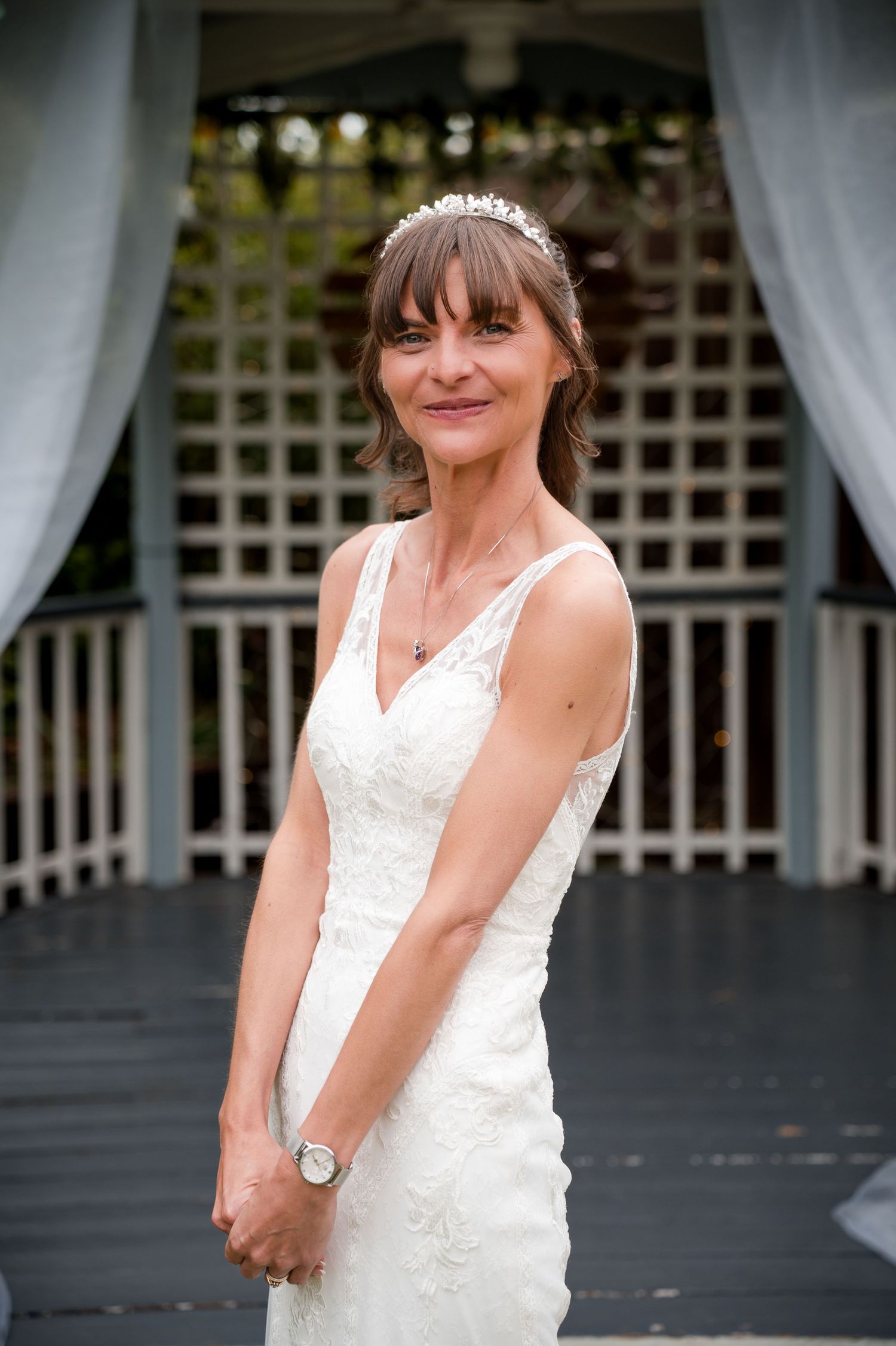 A bride in a white lace wedding dress poses outdoors near white curtains and lattice fencing.