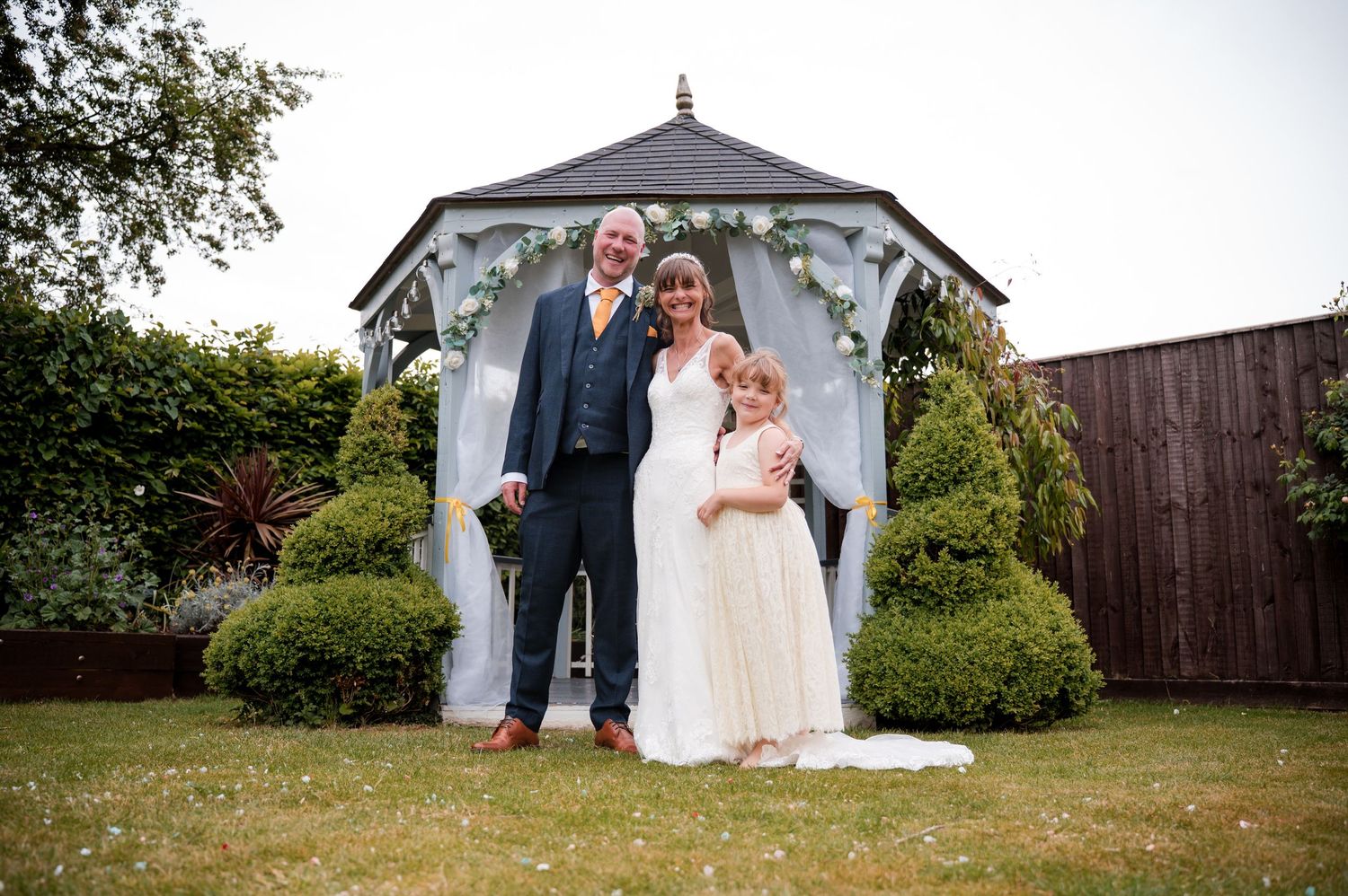 A couple stands under a white gazebo decorated with floral arrangements during an outdoor wedding ceremony.