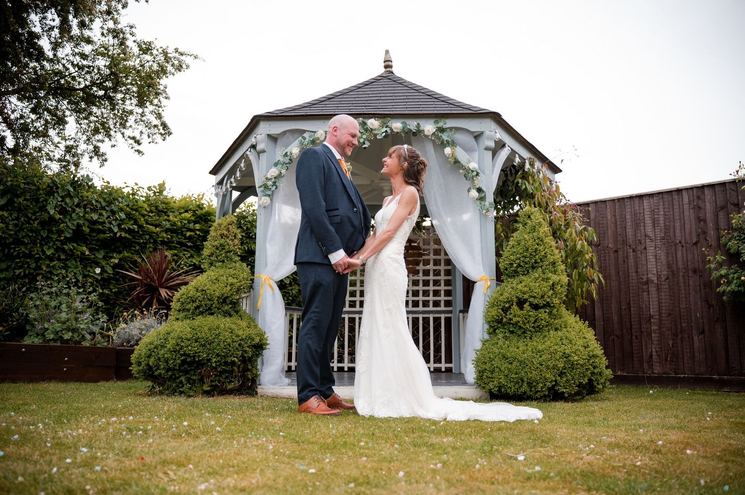 A romantic wedding couple shares a tender moment under a white gazebo in a garden setting.