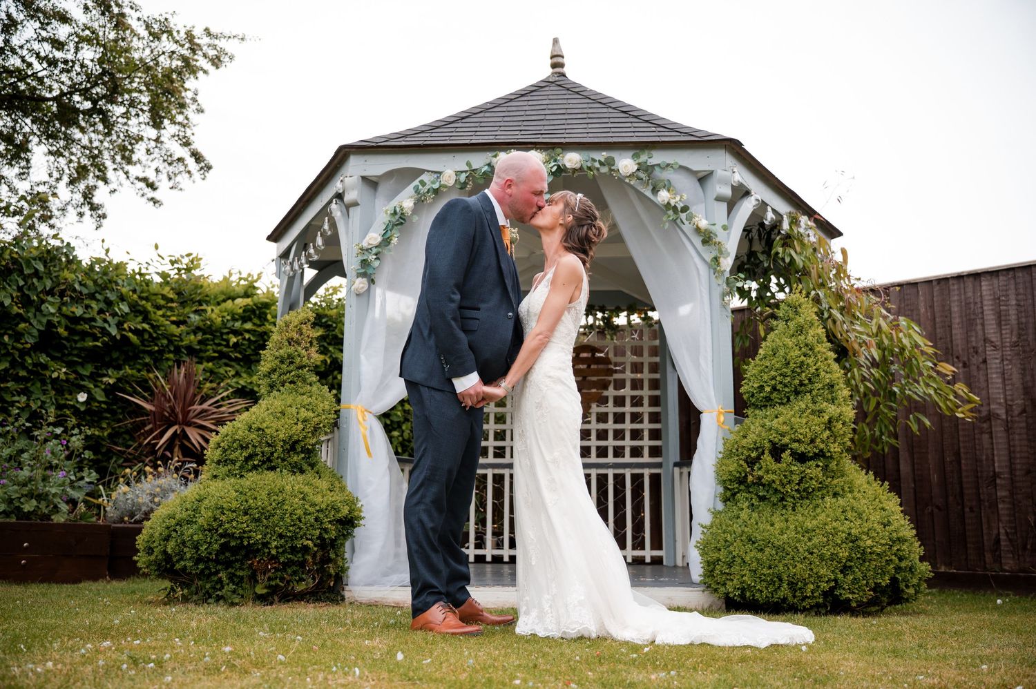 A romantic wedding couple shares a kiss in front of a white gazebo surrounded by lush garden greenery.