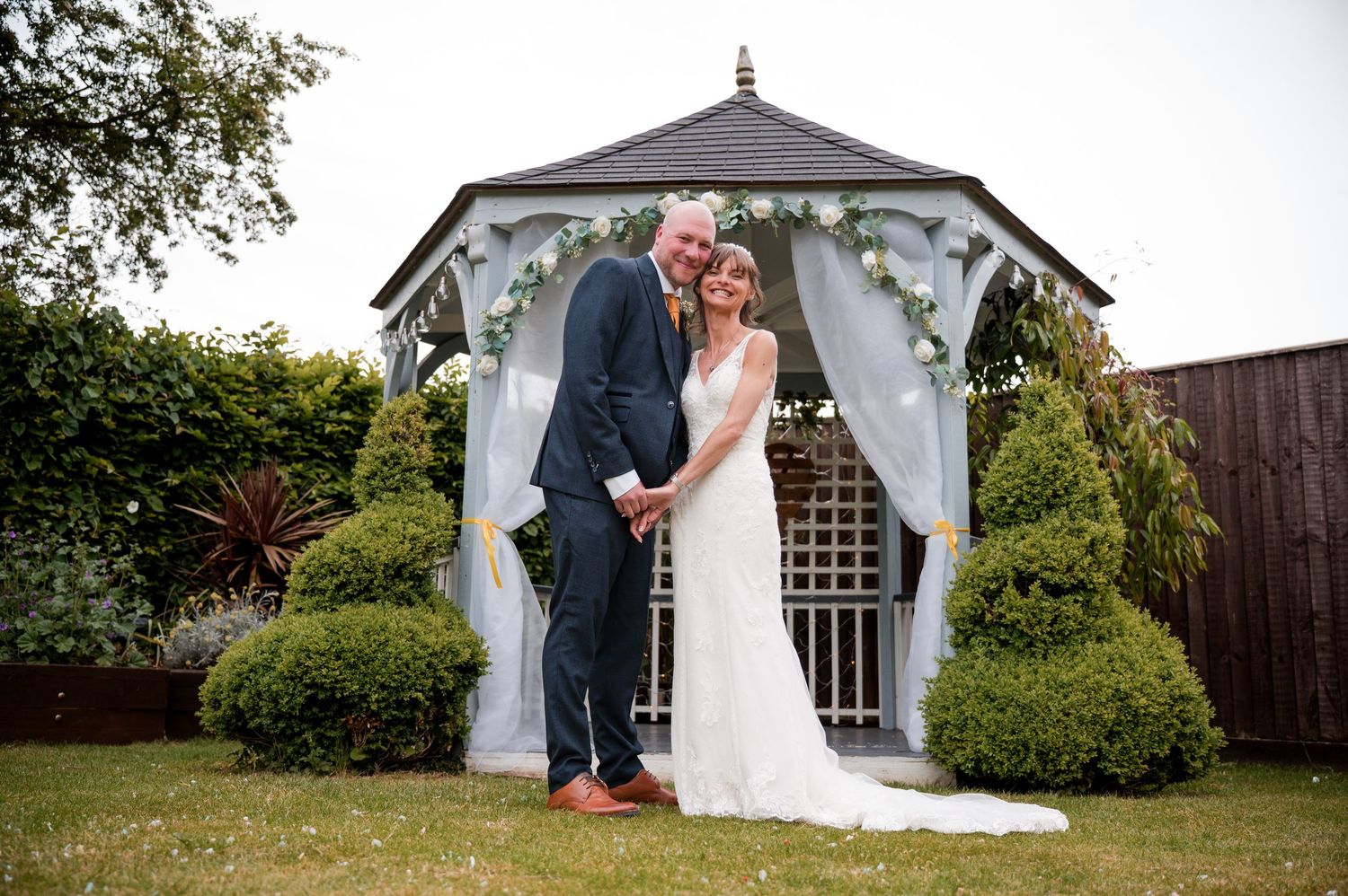 A romantic wedding ceremony scene in a garden gazebo with white floral decorations and elegant formal attire.
