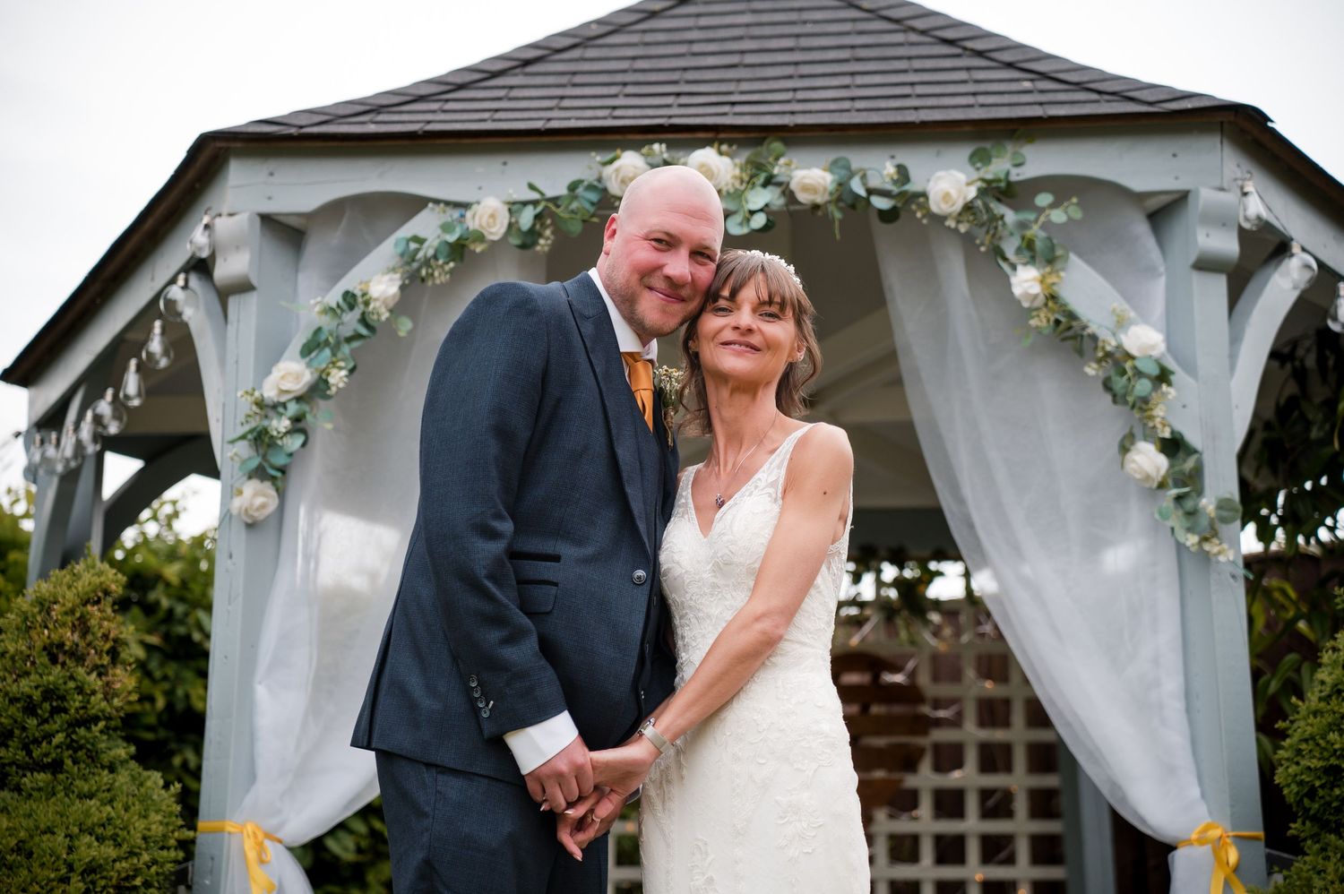 Newlywed couple shares a romantic moment under a white draped gazebo during an outdoor wedding ceremony.