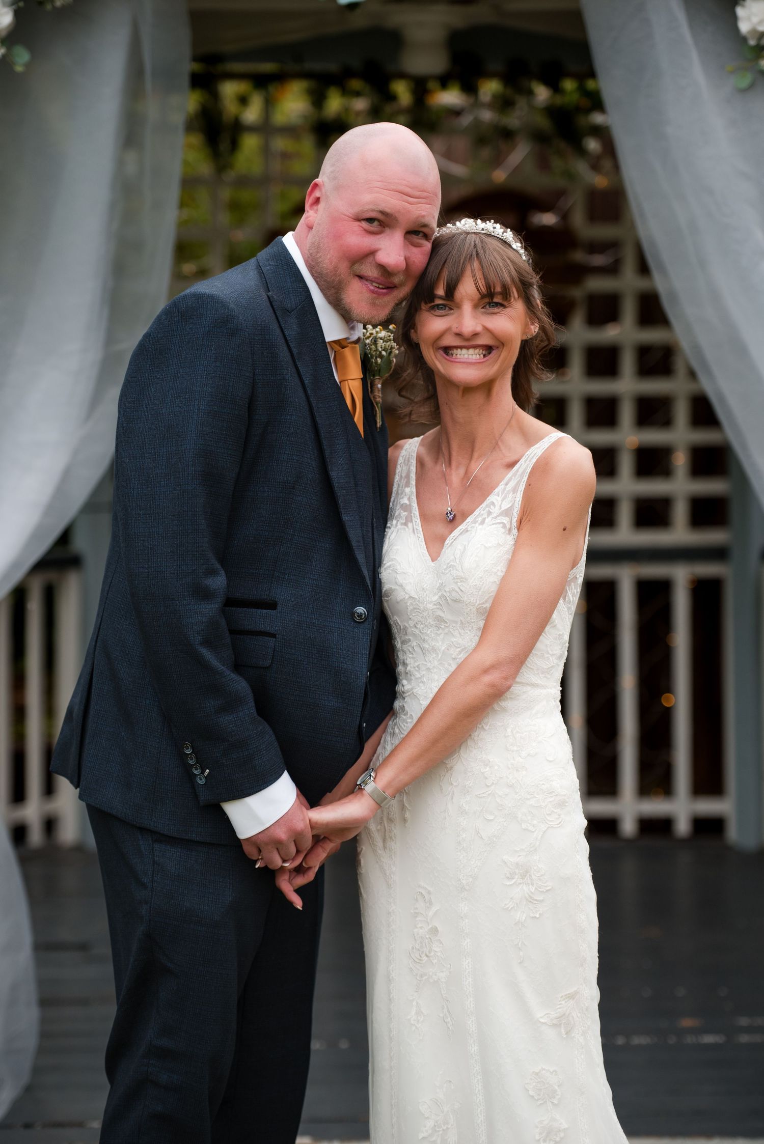 A couple shares a romantic moment in formal wedding attire at an outdoor venue with white curtains.