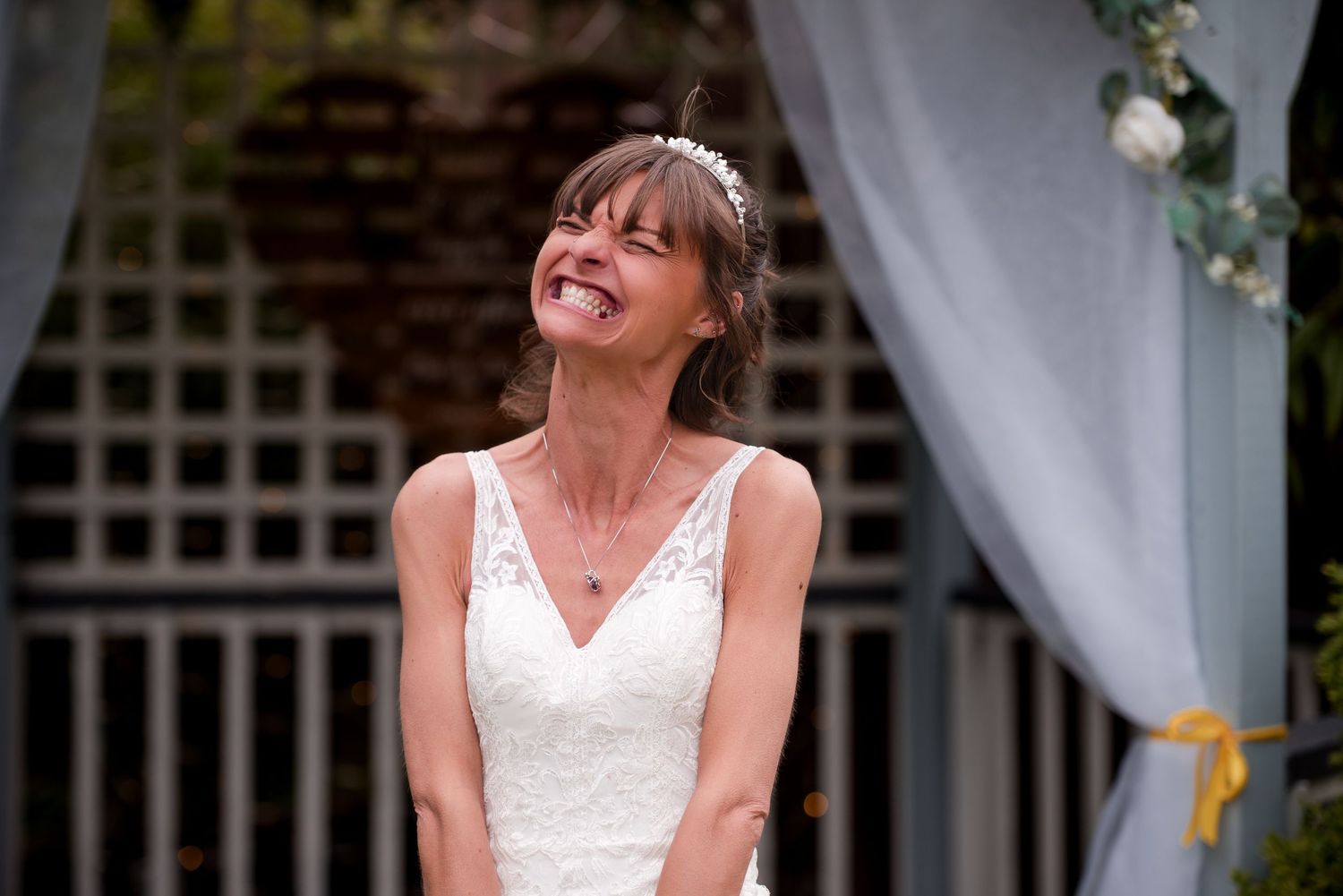 A joyful person in a white sleeveless dress laughs while posing against a decorative outdoor backdrop.