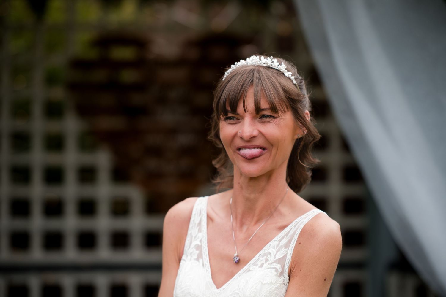 A bride in a white lace wedding dress smiles joyfully during an outdoor celebration.