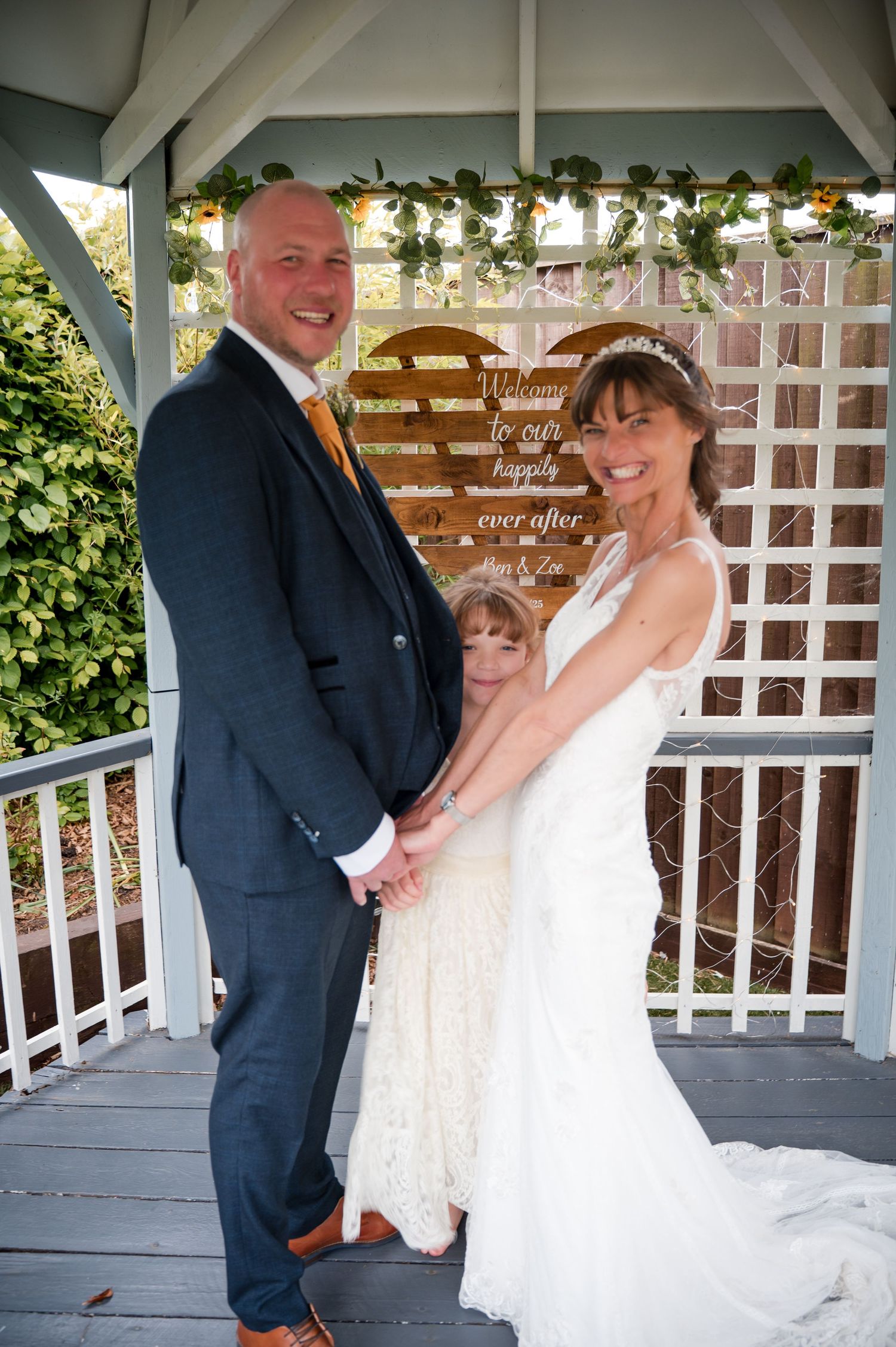 A wedding couple poses together in a white gazebo decorated with floral garlands and string lights.