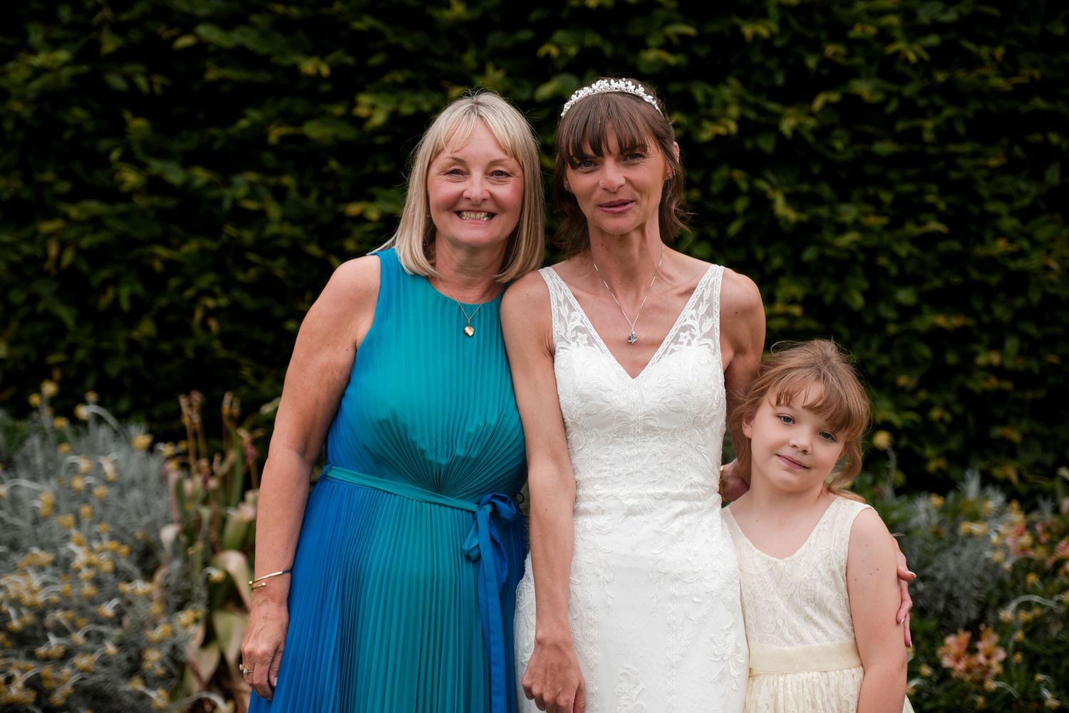 Three people wearing white and turquoise dresses pose together in an outdoor garden setting.