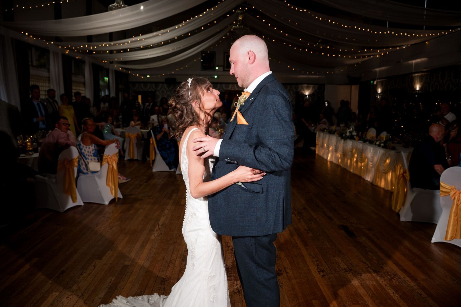 A couple shares a romantic dance together on a wooden floor beneath elegant draped lighting at a formal event.