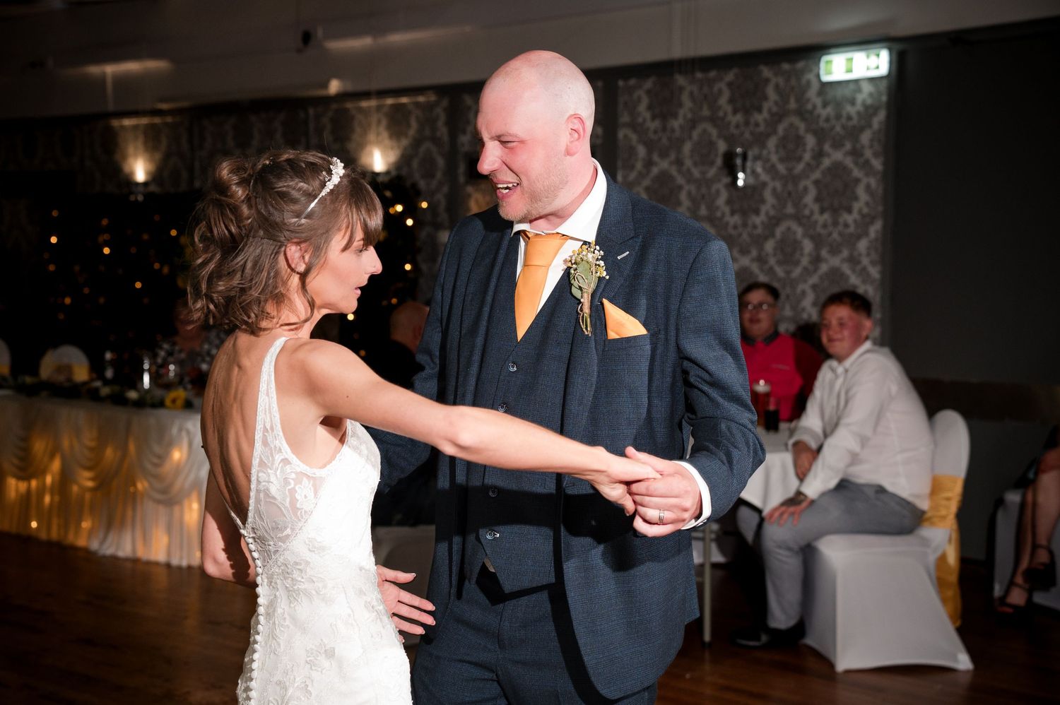 A couple shares a romantic dance together at an elegant wedding reception with ornate wallpaper in the background.