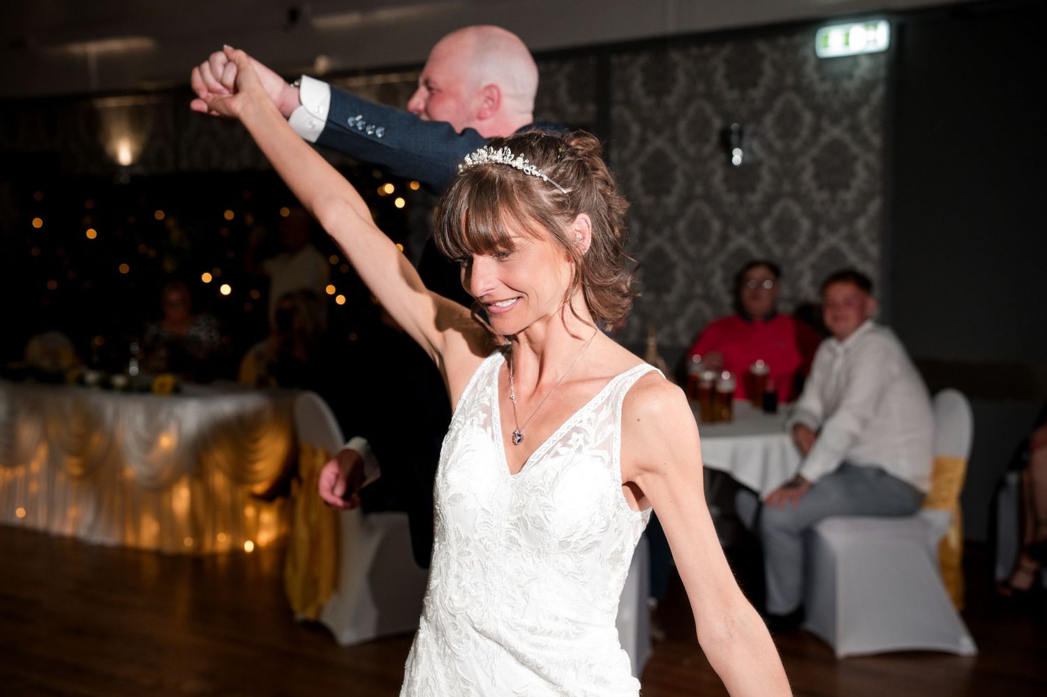 A couple dances together at a wedding reception with guests seated at tables in the background.