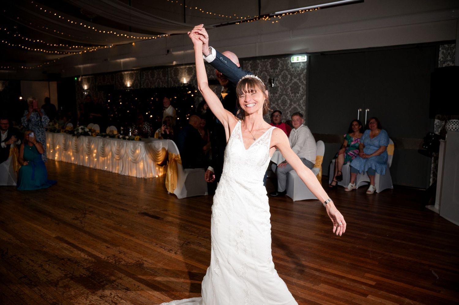 A joyful dancer in a white wedding dress twirls on a wooden dance floor under string lights at a reception.