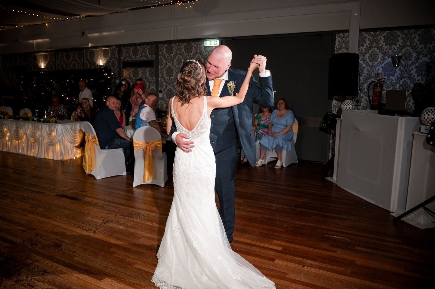 A couple shares a romantic dance on a wooden floor at a wedding reception with guests watching in the background.