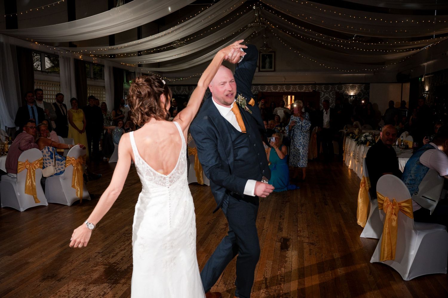 A couple dances together at an elegant wedding reception in a ballroom with draped decor and white chair covers.