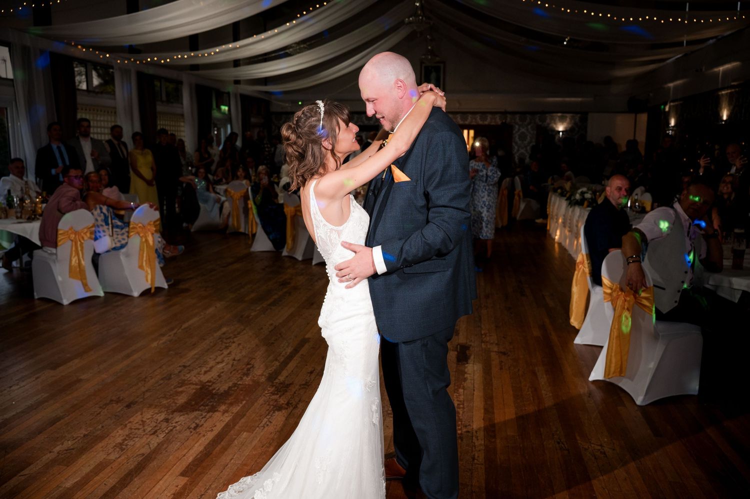 A couple shares their first dance at a wedding reception on a wooden dance floor under soft lighting.