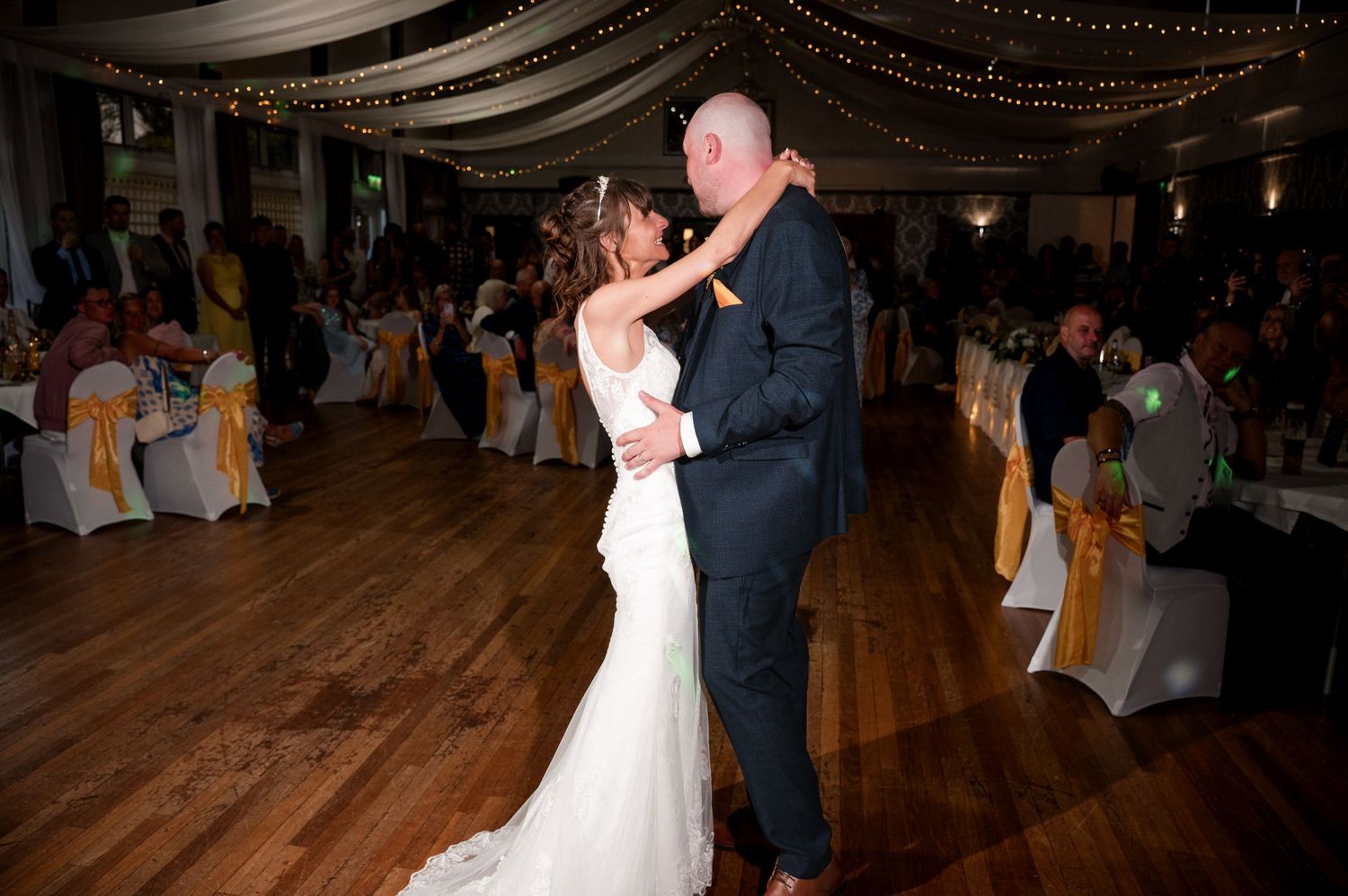 Couple dancing together at an elegant wedding reception with draped fabric decor and golden chair covers in the background.