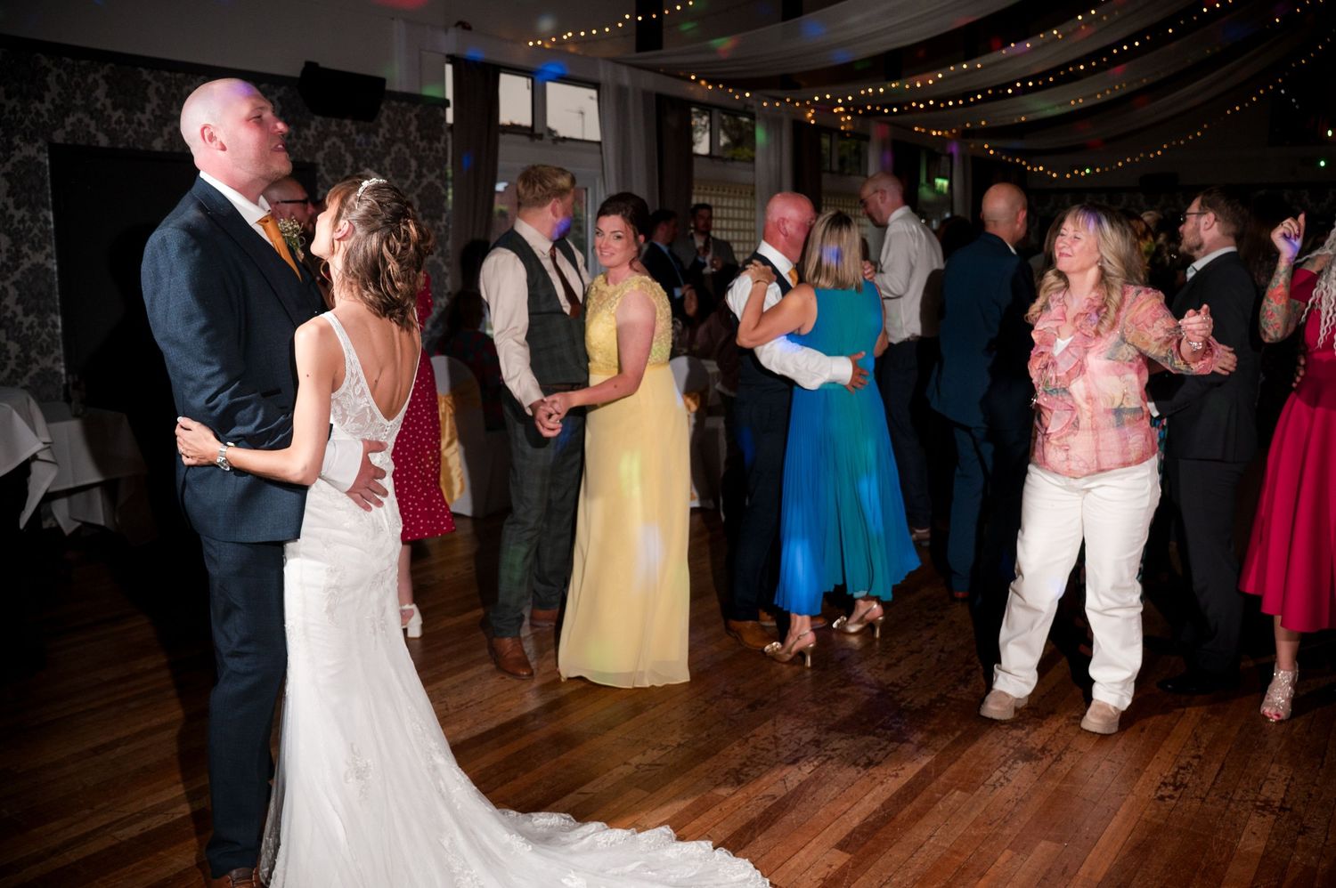 Wedding guests dance together on a wooden dance floor at an indoor reception venue with string lights overhead.