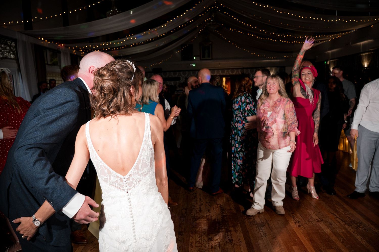 Wedding guests dancing and celebrating on a dimly lit dance floor with string lights overhead during an evening reception.