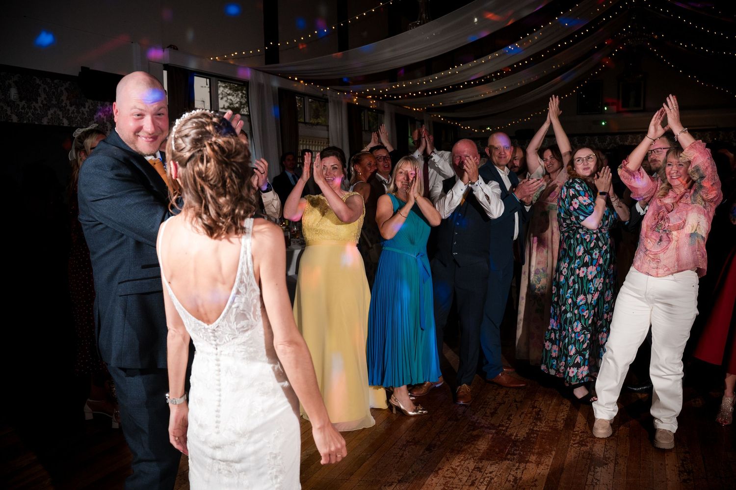 Wedding guests dance together on a dimly lit dance floor during an evening reception celebration.