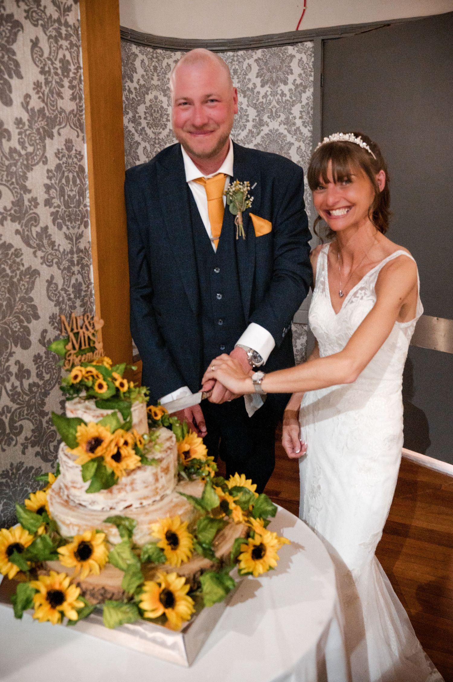 A wedding couple cuts their white tiered cake decorated with sunflowers at their reception.