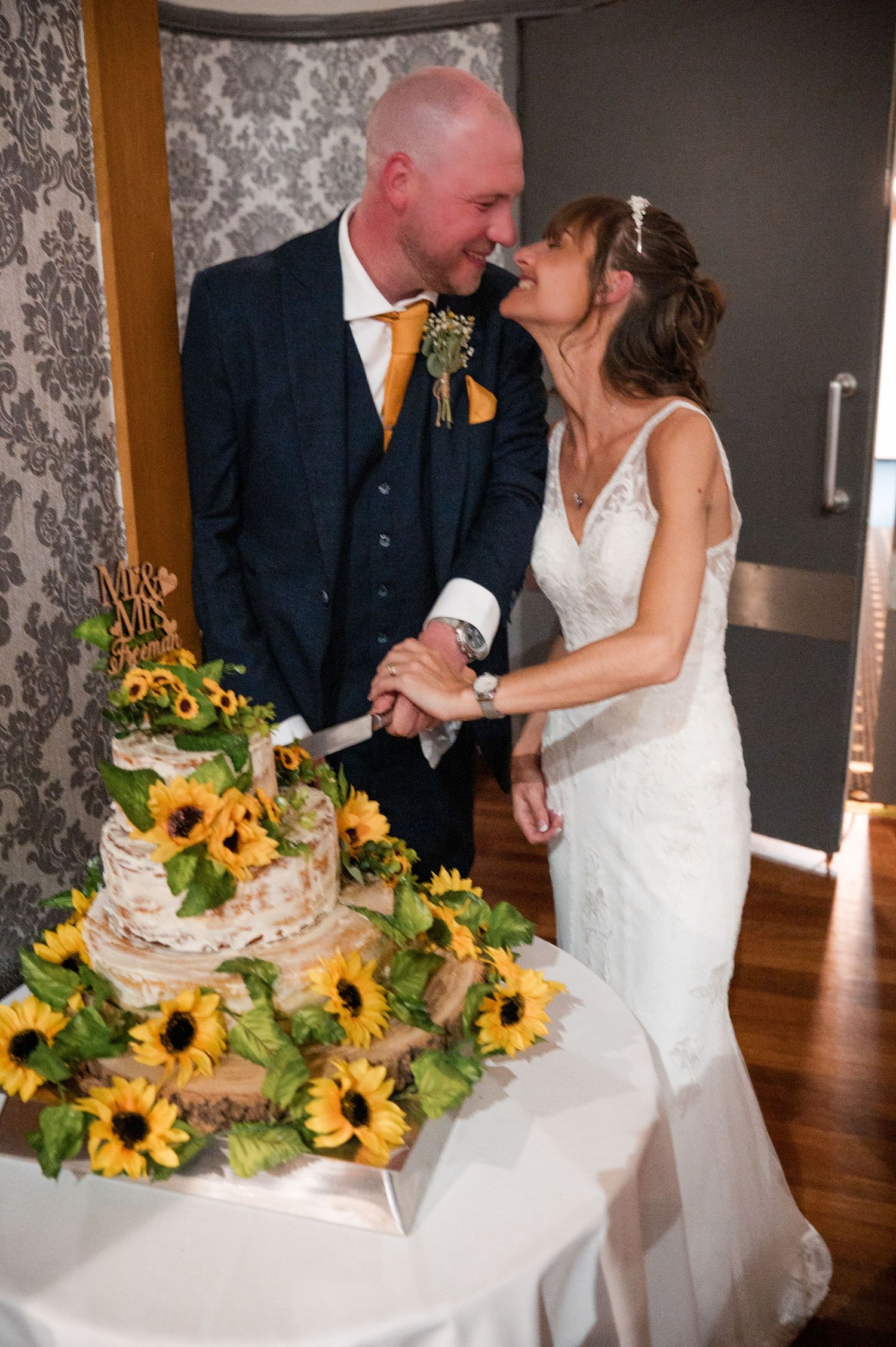 Wedding couple shares romantic moments while cutting their rustic cake decorated with vibrant sunflowers.