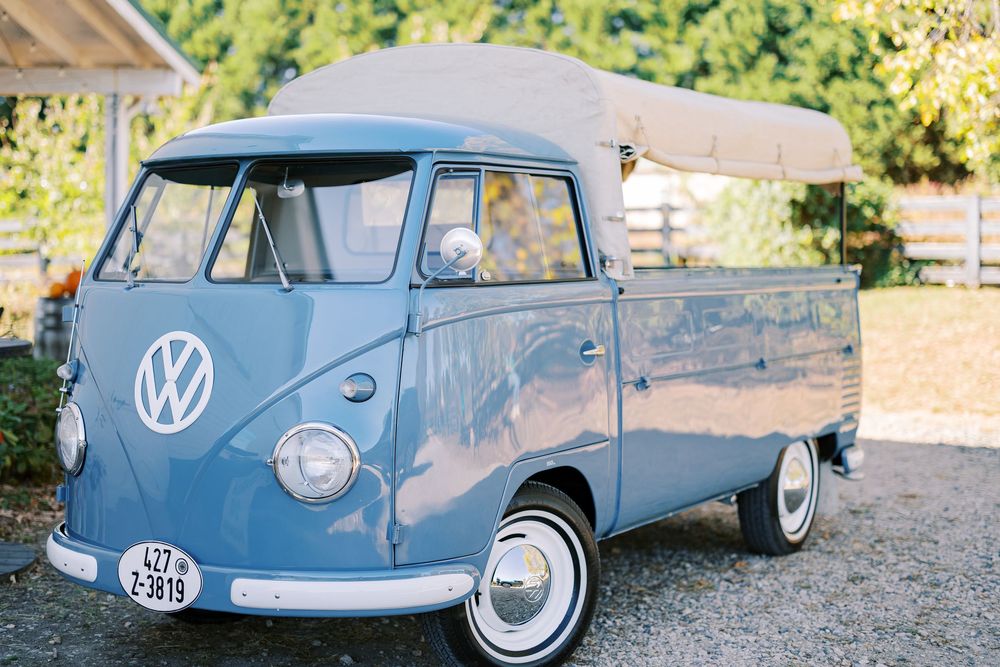 A vintage blue Volkswagen van parked on gravel with trees in the background on a sunny day.