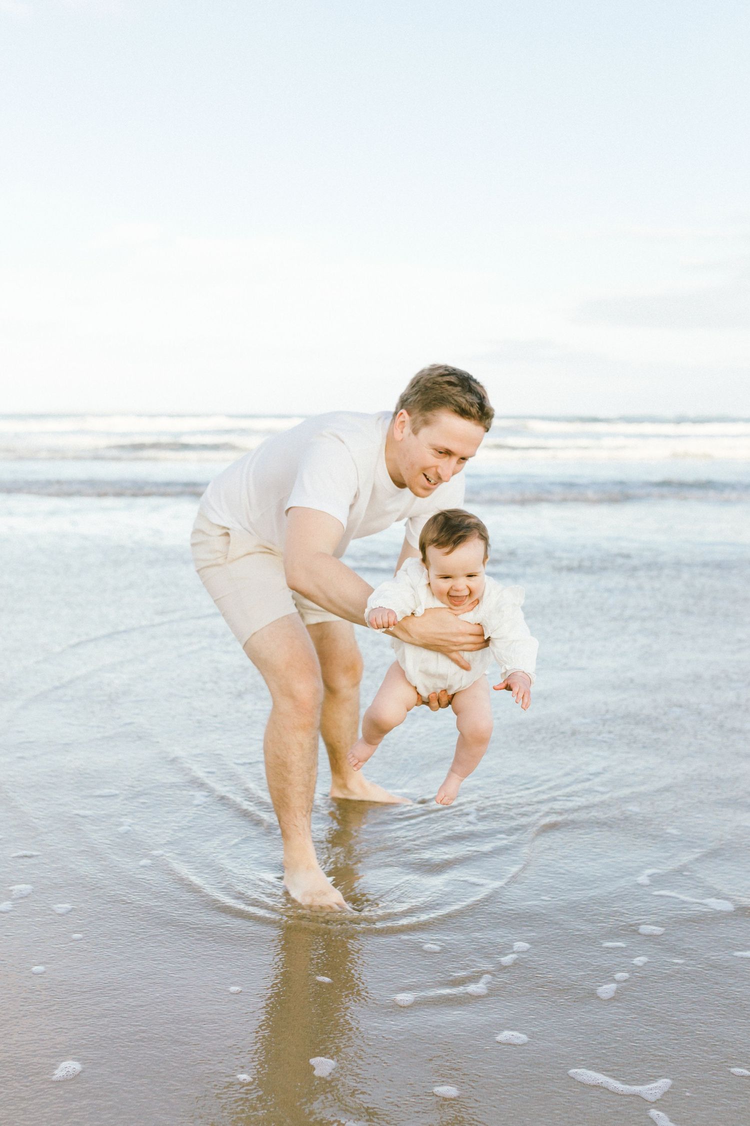Parent and baby play together at the edge of the ocean waves on a sandy beach.