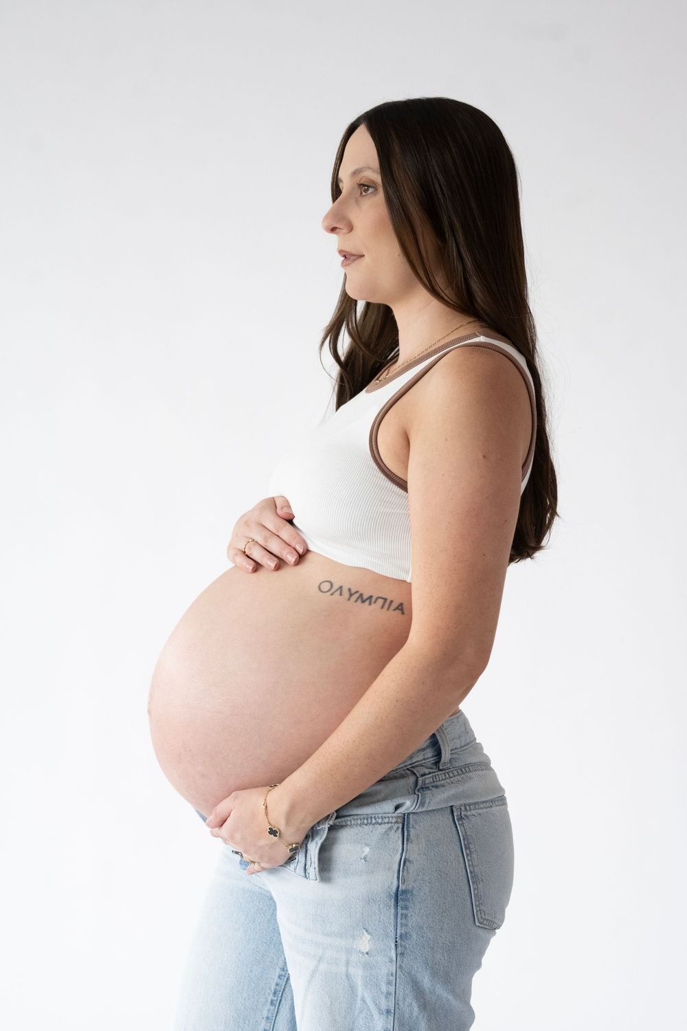 Side view of pregnant woman in white tank top and jeans holding her belly against white background.