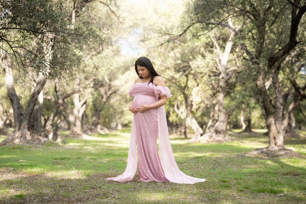 Expectant mother in flowing pink dress poses in olive grove during sunset maternity photoshoot.