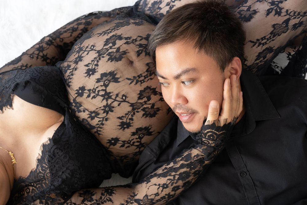 A man in a black shirt rests his head on his hand while lying next to a woman in black lace lingerie.