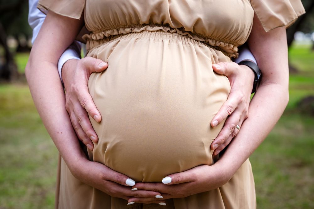 Pregnant woman in beige dress with partner's hands cradling baby bump outdoors.