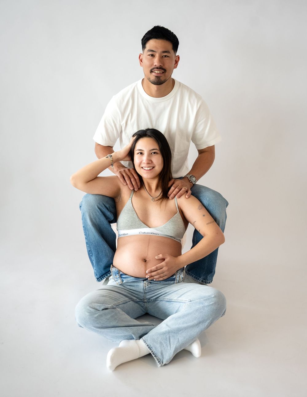 Couple maternity photo with pregnant woman sitting cross-legged on floor, partner behind her, both in jeans on white background.