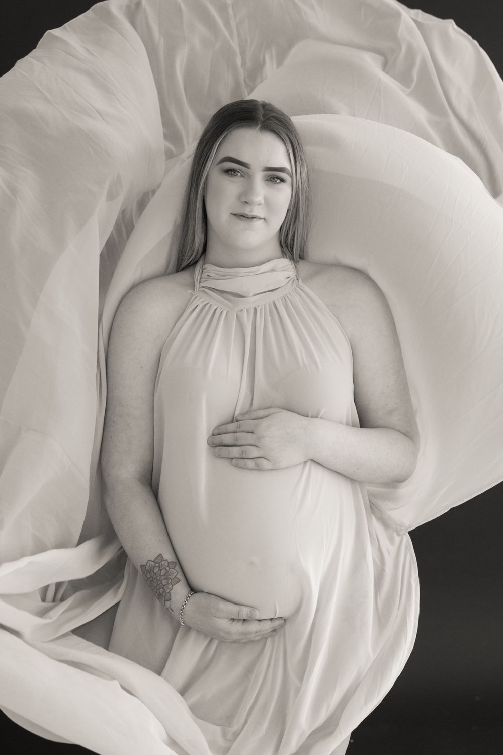 Black and white maternity photo of a pregnant woman in a flowing white halter dress, cradling her baby bump.