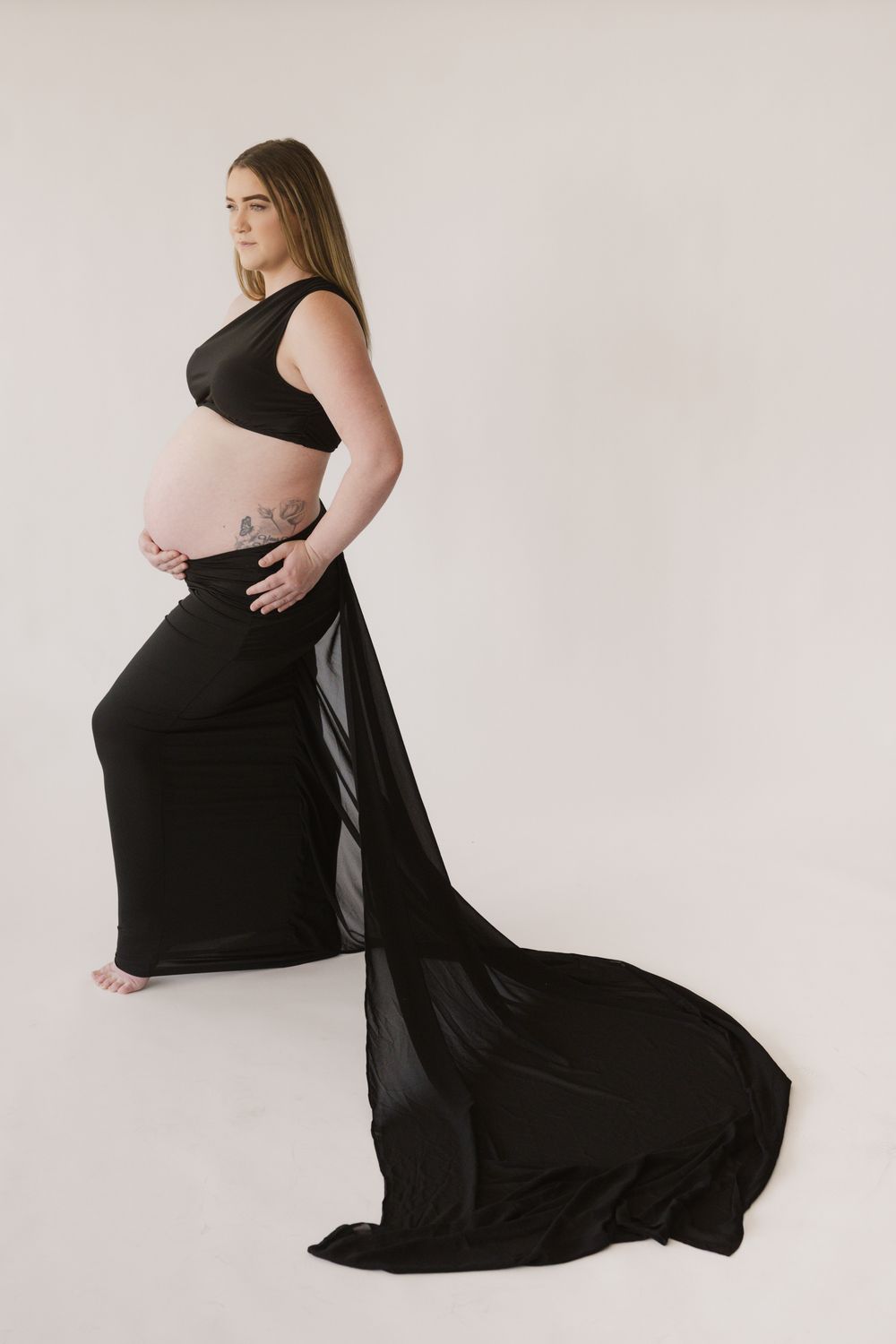 Pregnant woman in black two-piece maternity gown with flowing train poses against white background.