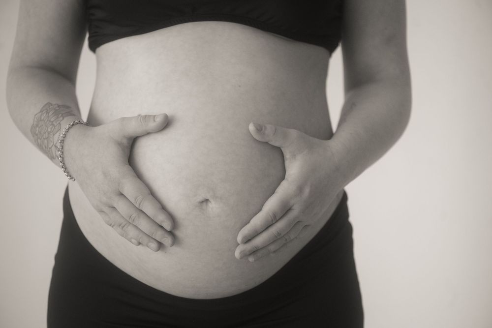 Black and white photo of a pregnant woman cradling her bare baby bump with both hands, wearing a black top.