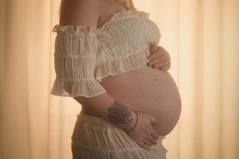 Pregnant woman in white ruffled off-shoulder dress cradling bare baby bump, with floral wrist tattoo, soft golden light.
