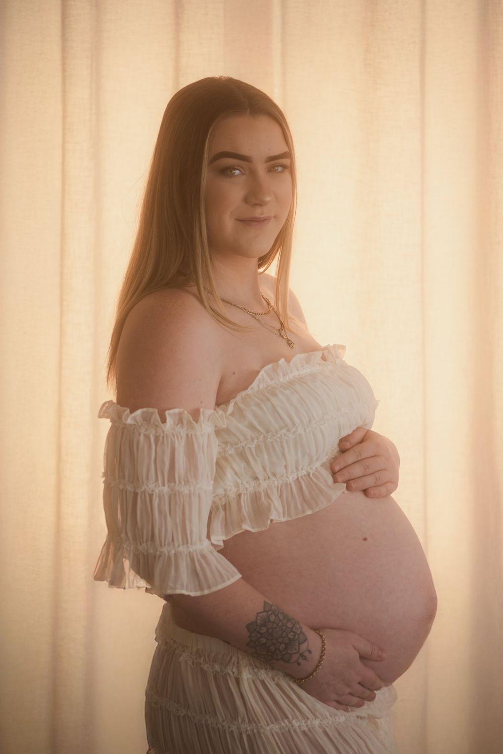 Pregnant woman in white off-shoulder ruffled top cradling her baby bump, glowing in warm backlit light.