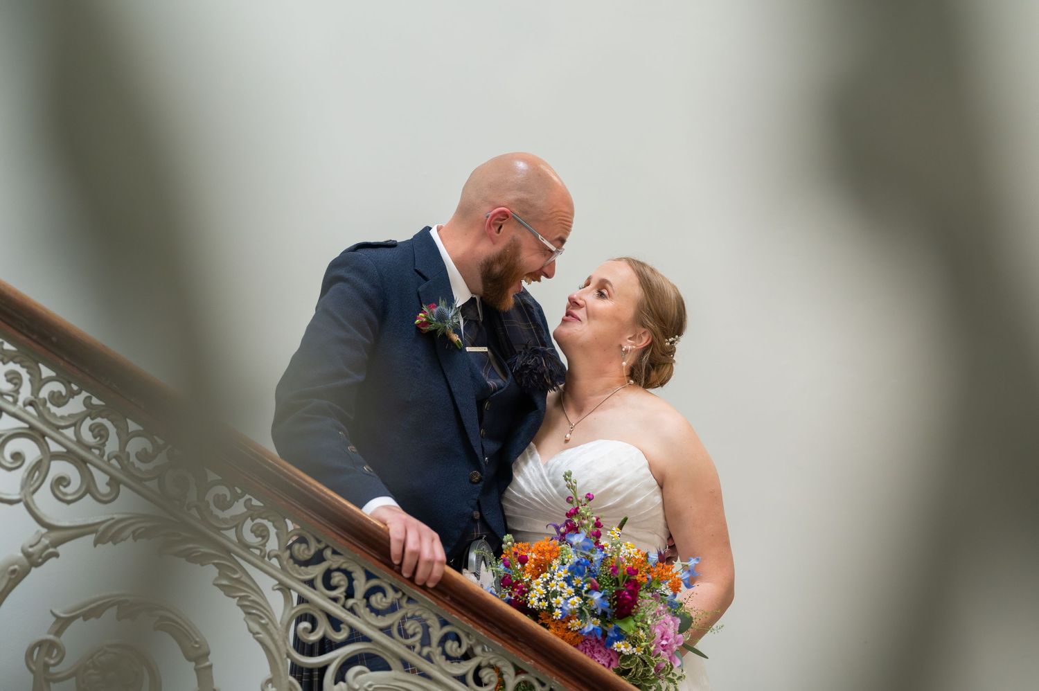 A couple shares a romantic kiss on an elegant staircase with ornate railings while holding a colorful bouquet.