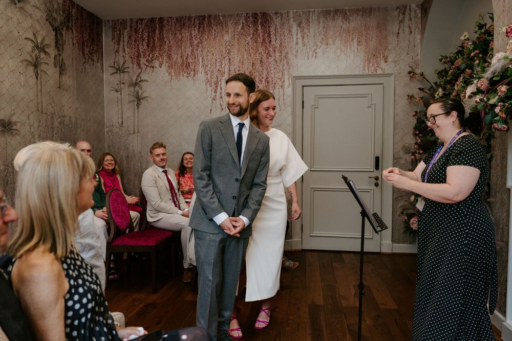 Couple smiling during intimate wedding ceremony in elegant floral venue with seated guests.