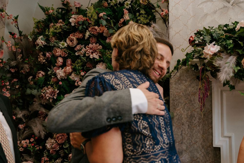 Two people share a warm embrace in front of a lush floral wall at a wedding celebration.