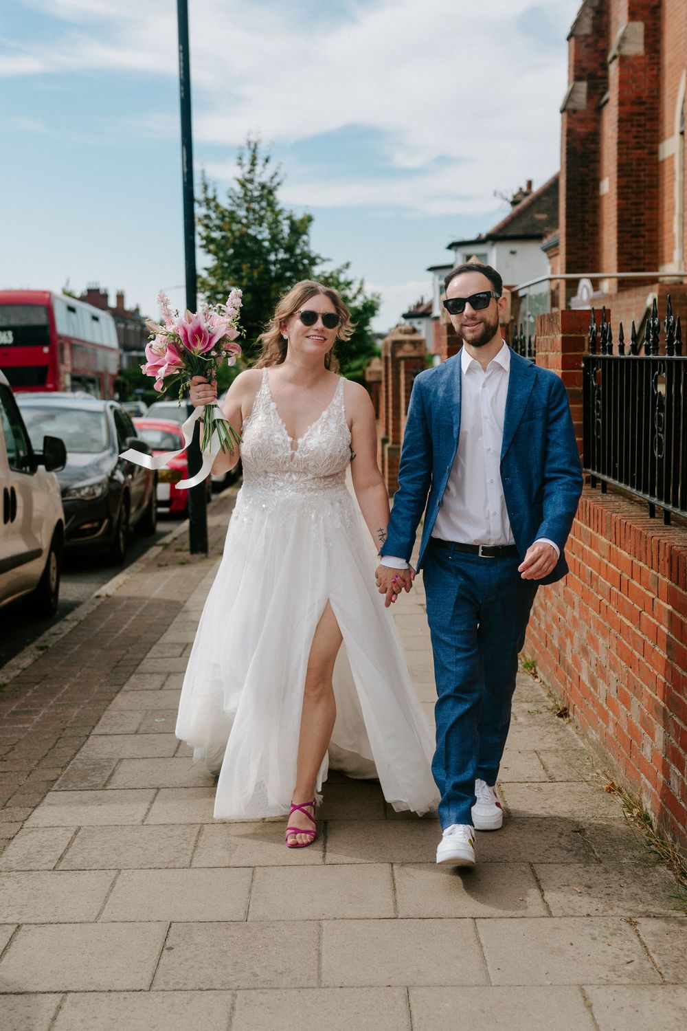 Newlywed couple walking down a sidewalk together on a sunny day with brick buildings in the background.