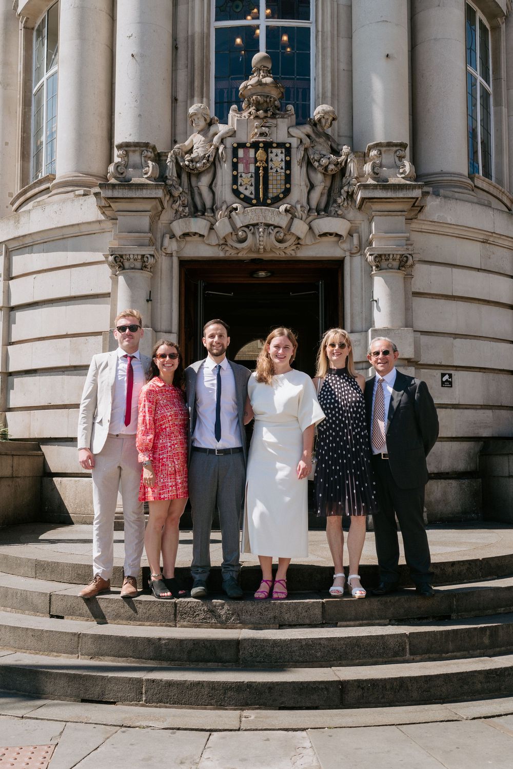 Group of six people posing on steps outside a grand neoclassical building on a sunny day.